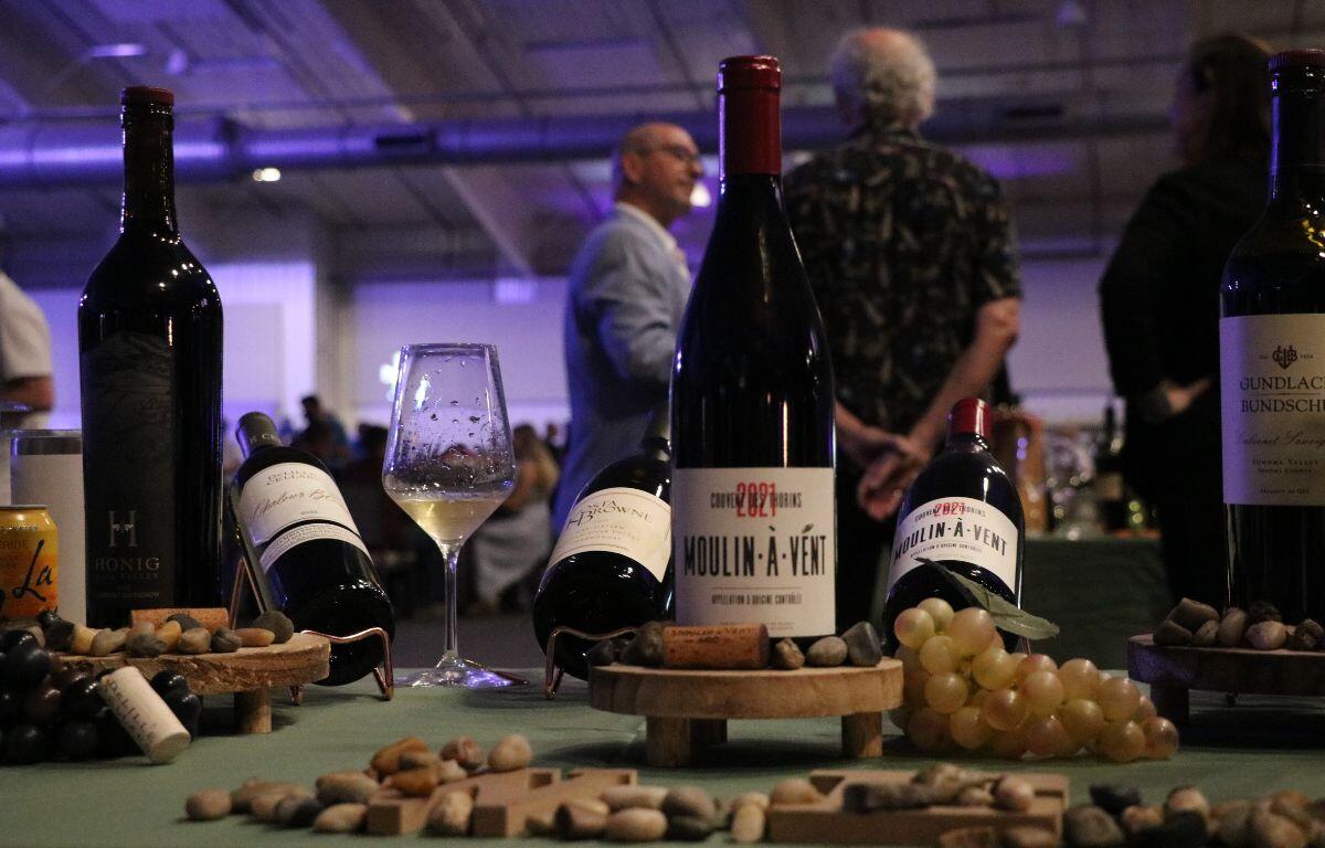 A table littered in rocks, fake grapes, a corkscrew and various alcohol bottles features a wine glass with alcohol in it. In the background are groups of people in a venue.