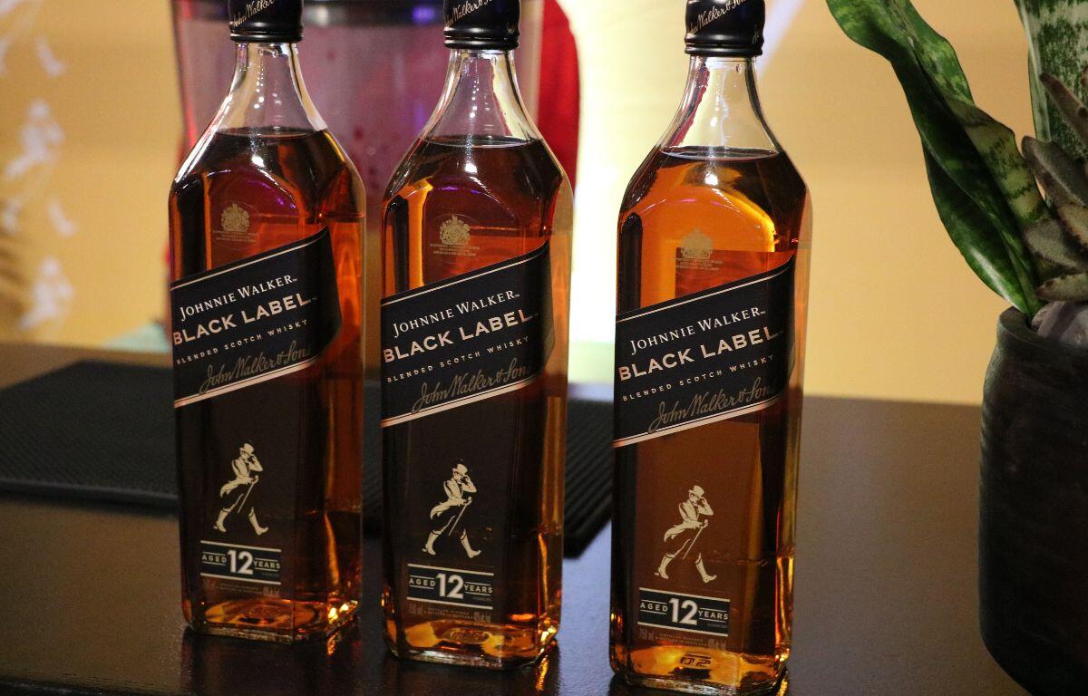 Three alcoholic beverages are lined up on a table with labels reading, "Johnnie Walker. Black Label." A plant is to the right of the bottles.