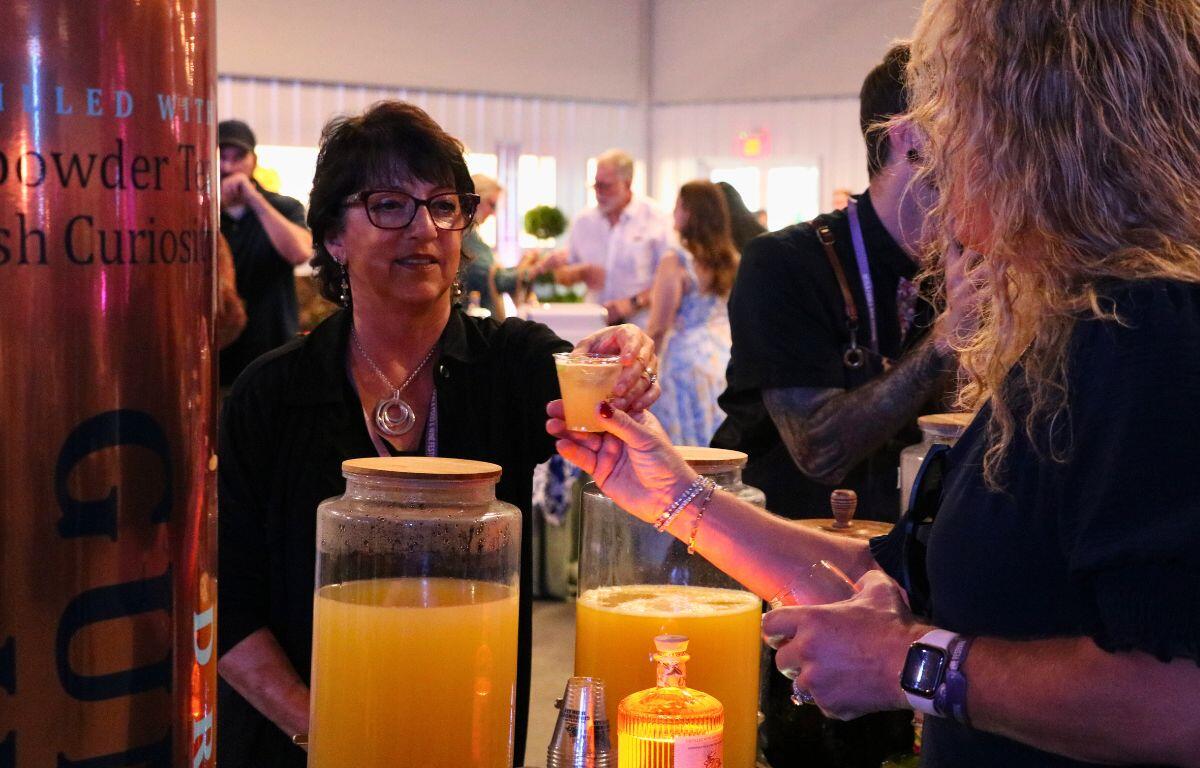 A woman stands behind a counter and hands an alcoholic beverage to another woman. Drinks are lined up on the table in front of her.