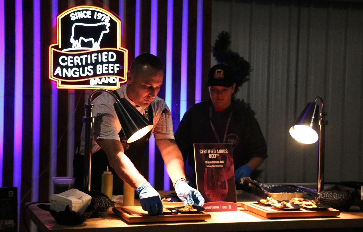 Two servers stand behind a wooden table with lights shining down onto small displays with samples of food on them. One of the servers places two sample plates onto the display. In the background is a neon light up sign reading "Since 1978. Certified Angus Beef Brand."