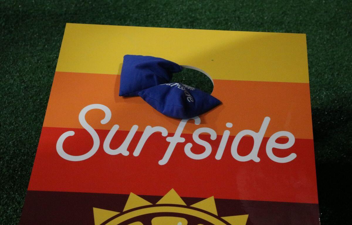 A yellow, orange, red and brown bean bag board has two blue bean bags with white text hanging from the board into the hole. The white text on the bean bags and the board read, "Surfside."