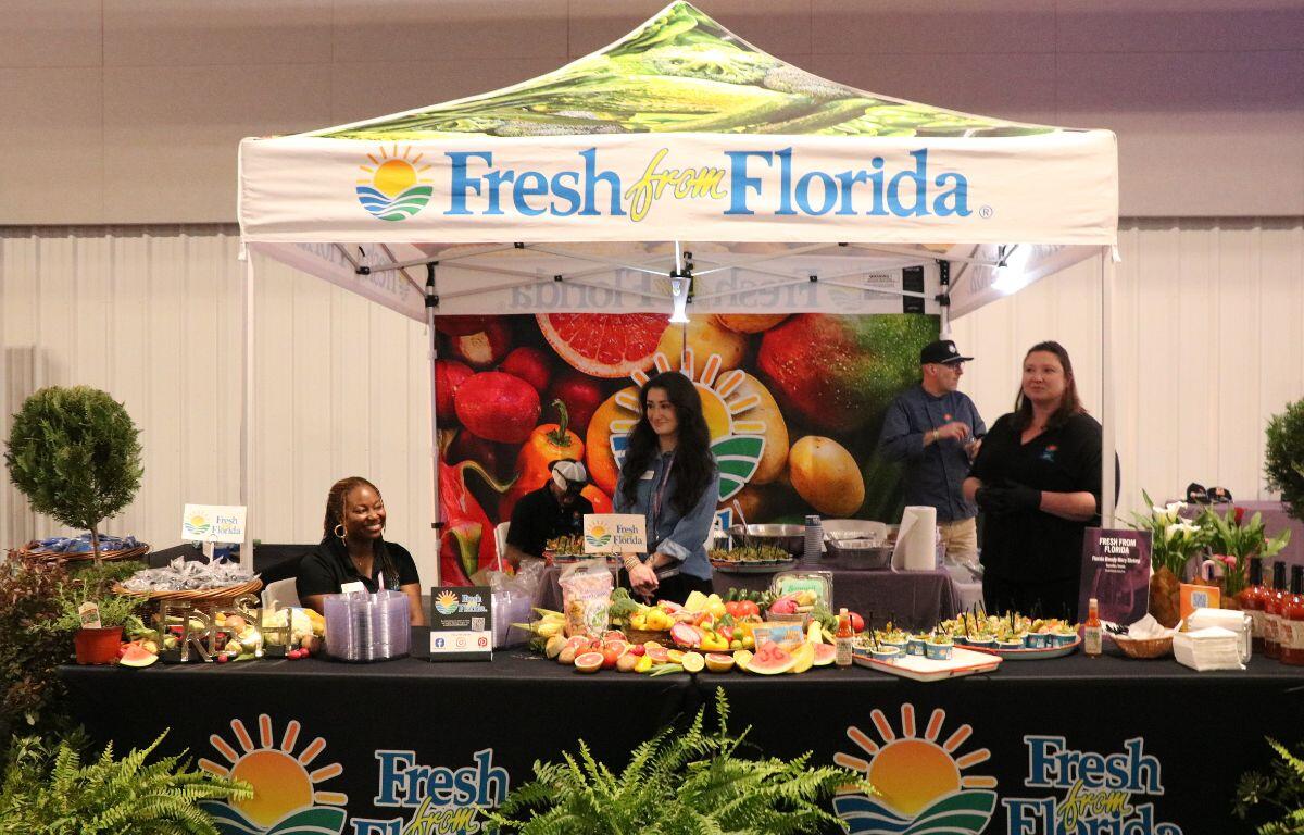 A canopy stands over a group of people behind a long table featuring fruits, shrimp, plants, sauce and more. The tent and table both have logos that read, "Fresh from Florida."
