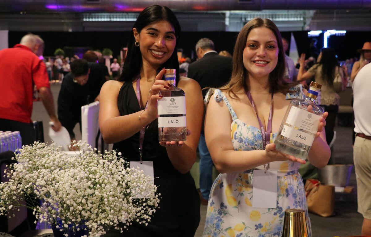 Two women hold alcoholic beverages with labels reading, "Lalo." In the background is a crowd of people walking around a venue.