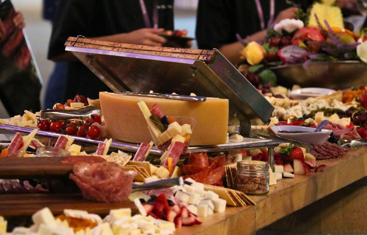 A long wooden table has a variety of fruits, cheese, vegetables, meats and other goods.