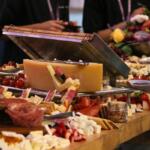 A long wooden table has a variety of fruits, cheese, vegetables, meats and other goods.