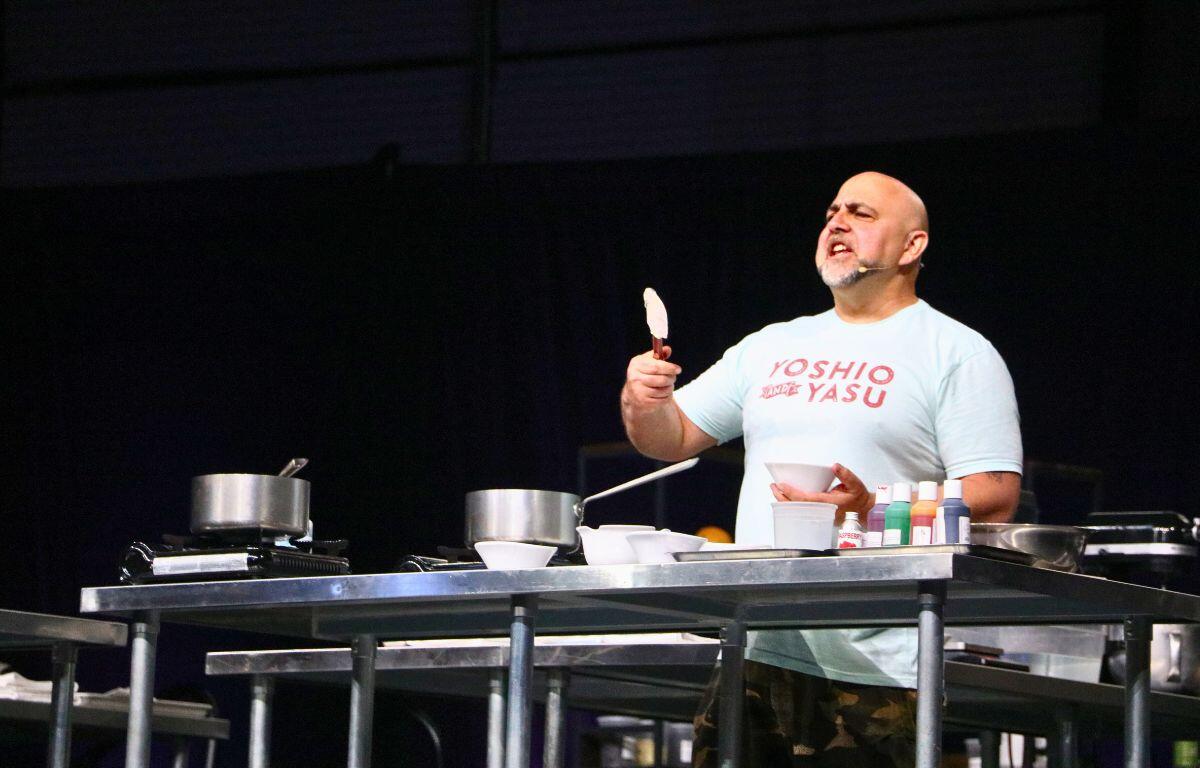 A man in a white shirt holds a cooking utensil at a cooking station engaging with an audience.