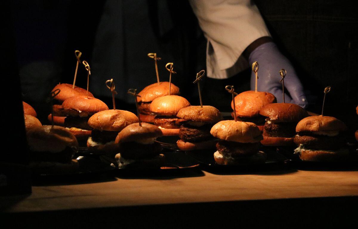 Sliders are lined up on a wooden table with sticks poking out of them to keep their shapes. Gloved hands push more sliders on the display.