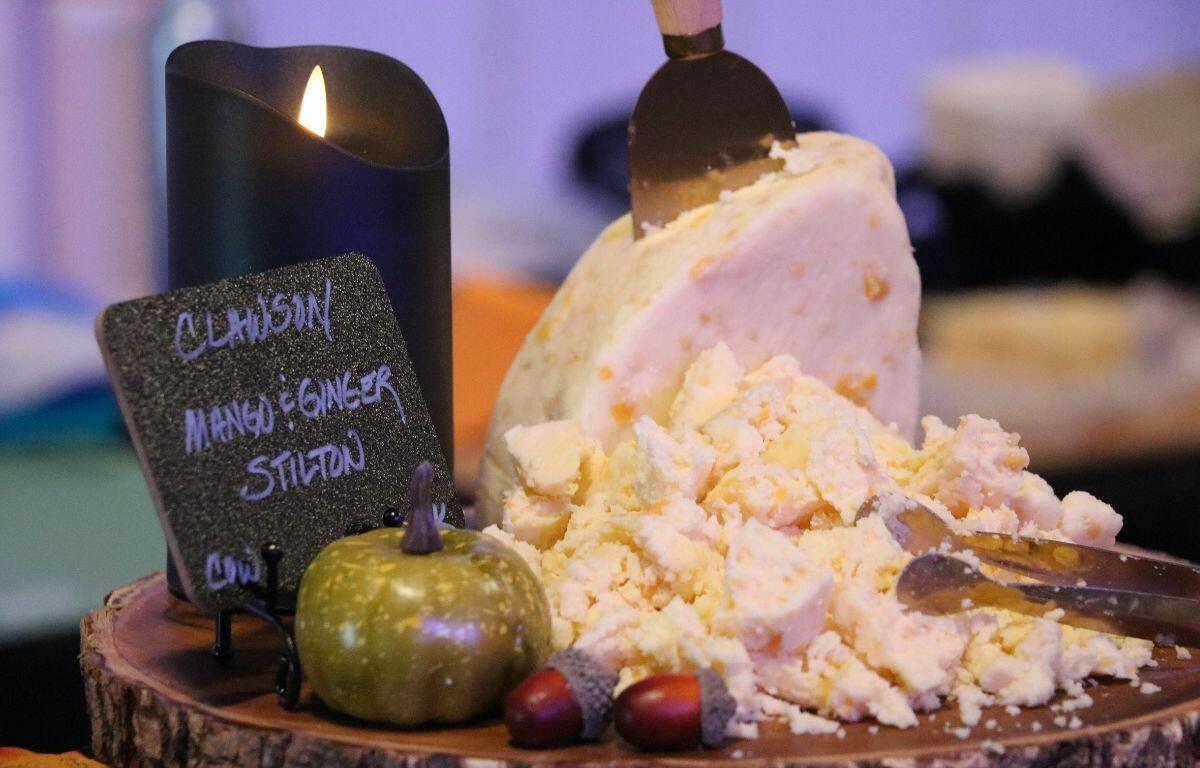 A piece of cheese with multiple shreds of cheese in front of it. There is a lit black candle to the left of the cheese and small decorations on the wood log displaying the cheese. There is a sign that reads, "Clawson Mango & Ginger Silton."