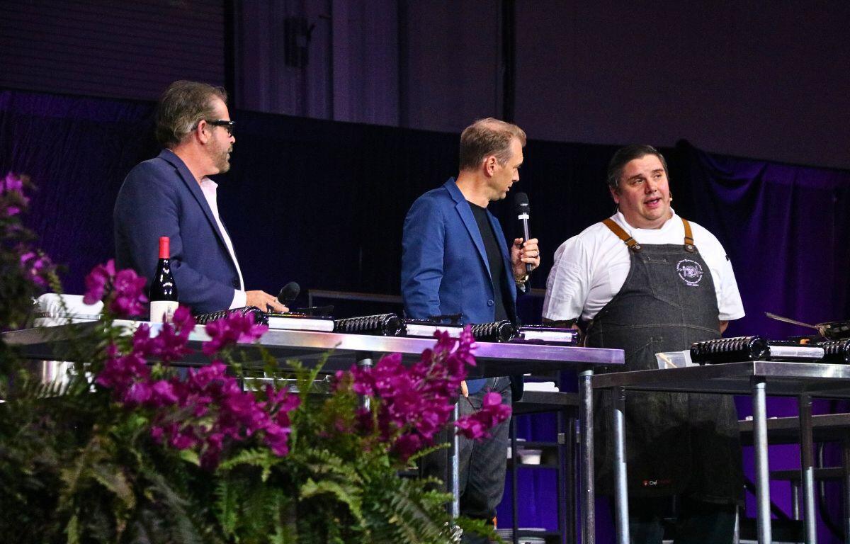 Three men gather around a metal table with cooking equipment. One man holds a microphone with purple flowers in the foreground.