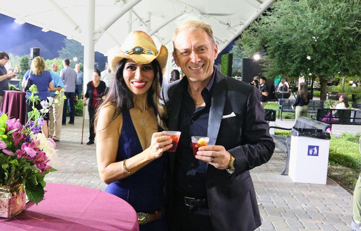 A man and a woman hold cocktails huddled together at a draped table in an outdoor venue.