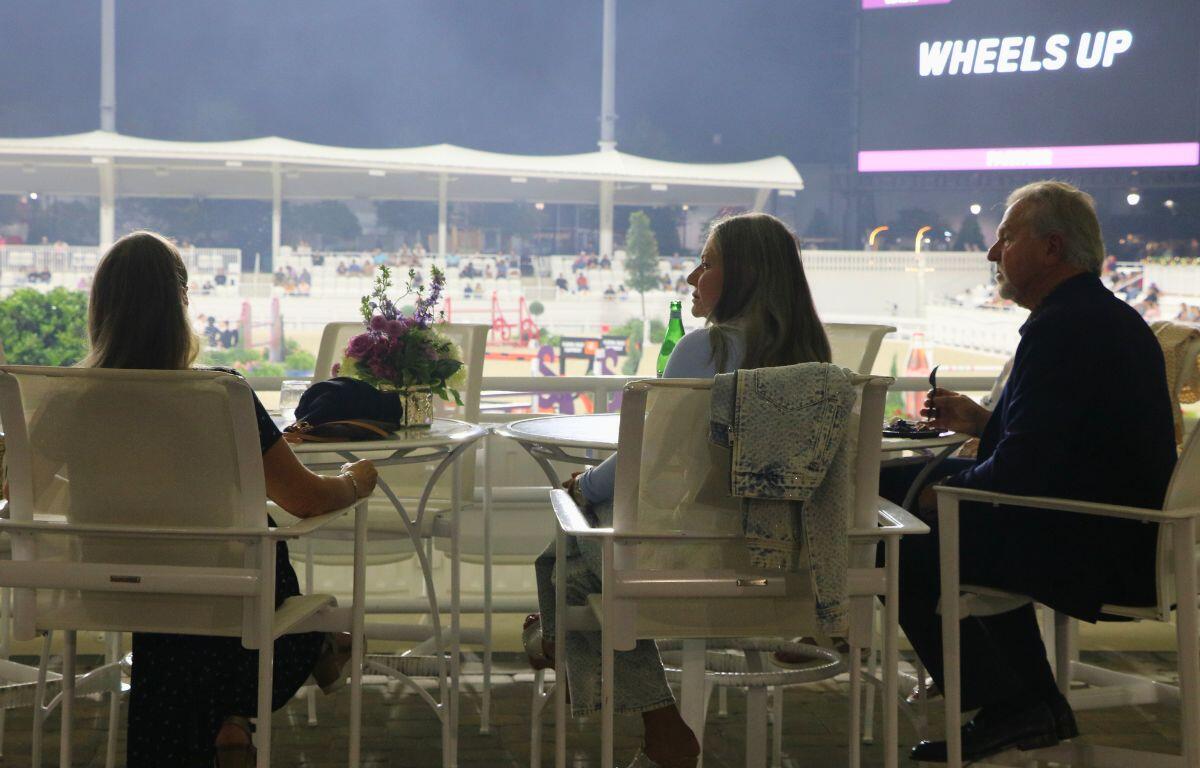 Three people sit in white chairs in front of tables with a large stadium in the background.