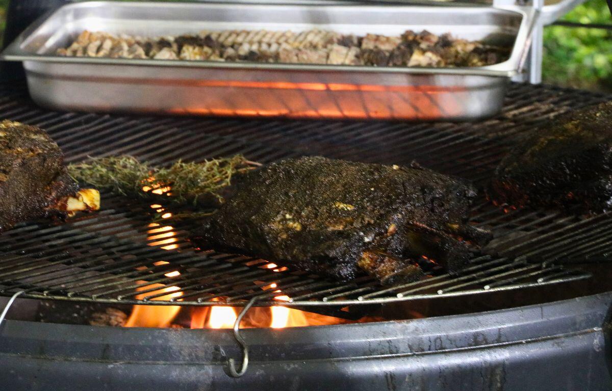 Large pieces of dark meat lay on a burner over an open fire. A tray of more meat sit in a tin behind it.