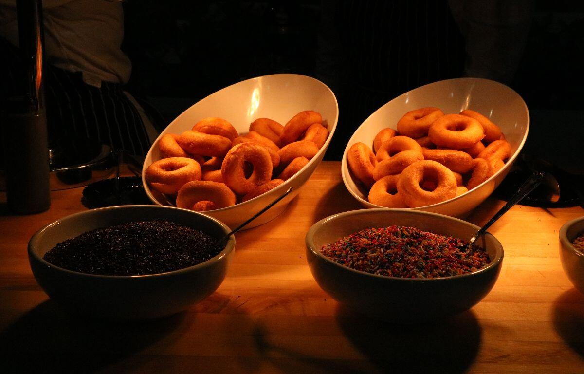 Two white, circular bowls of plane donuts sit under a bright light. A bowl of chocolate sprinkles sits on the left and a bowl of colorful sprinkles sits on the right.