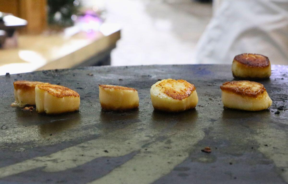 Golden scallops sit on a burner with people in chef's outfits in the background.