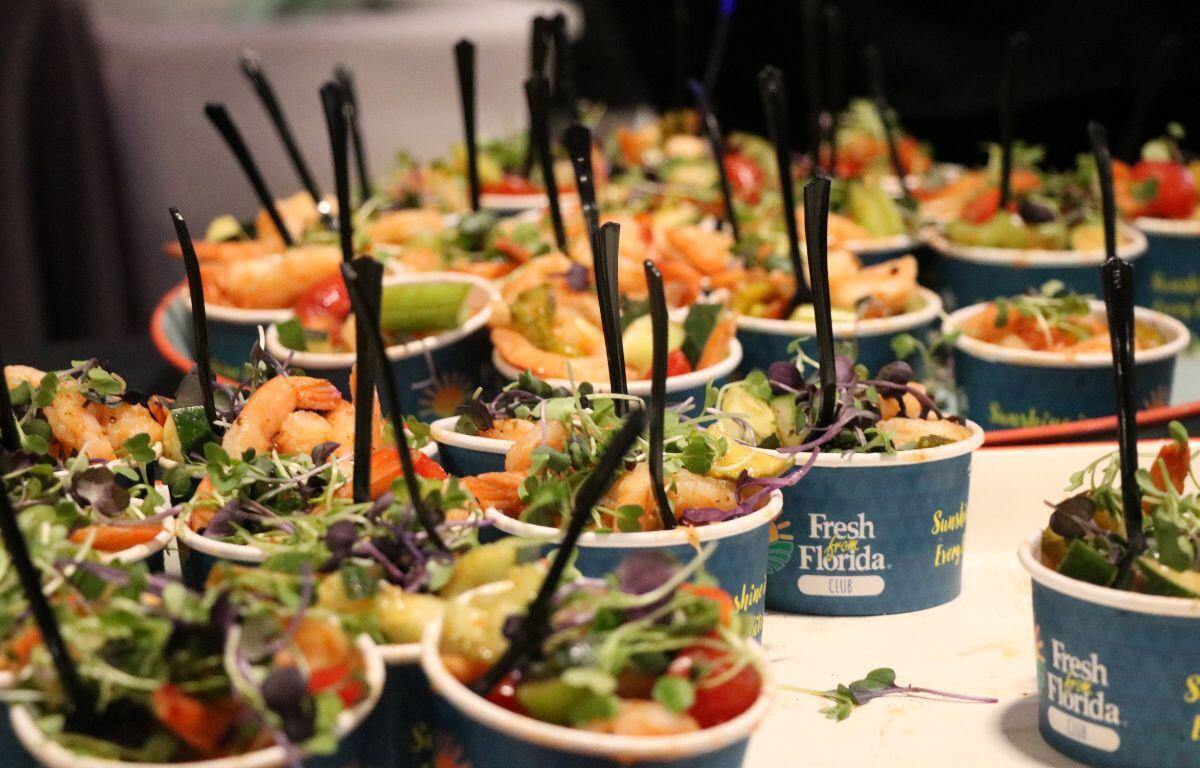 Multiple serving sizes of shrimp salads are lined up on a table with black cutlery sticking out of them. The containers are blue with "Fresh from Florida Club" printed on them.