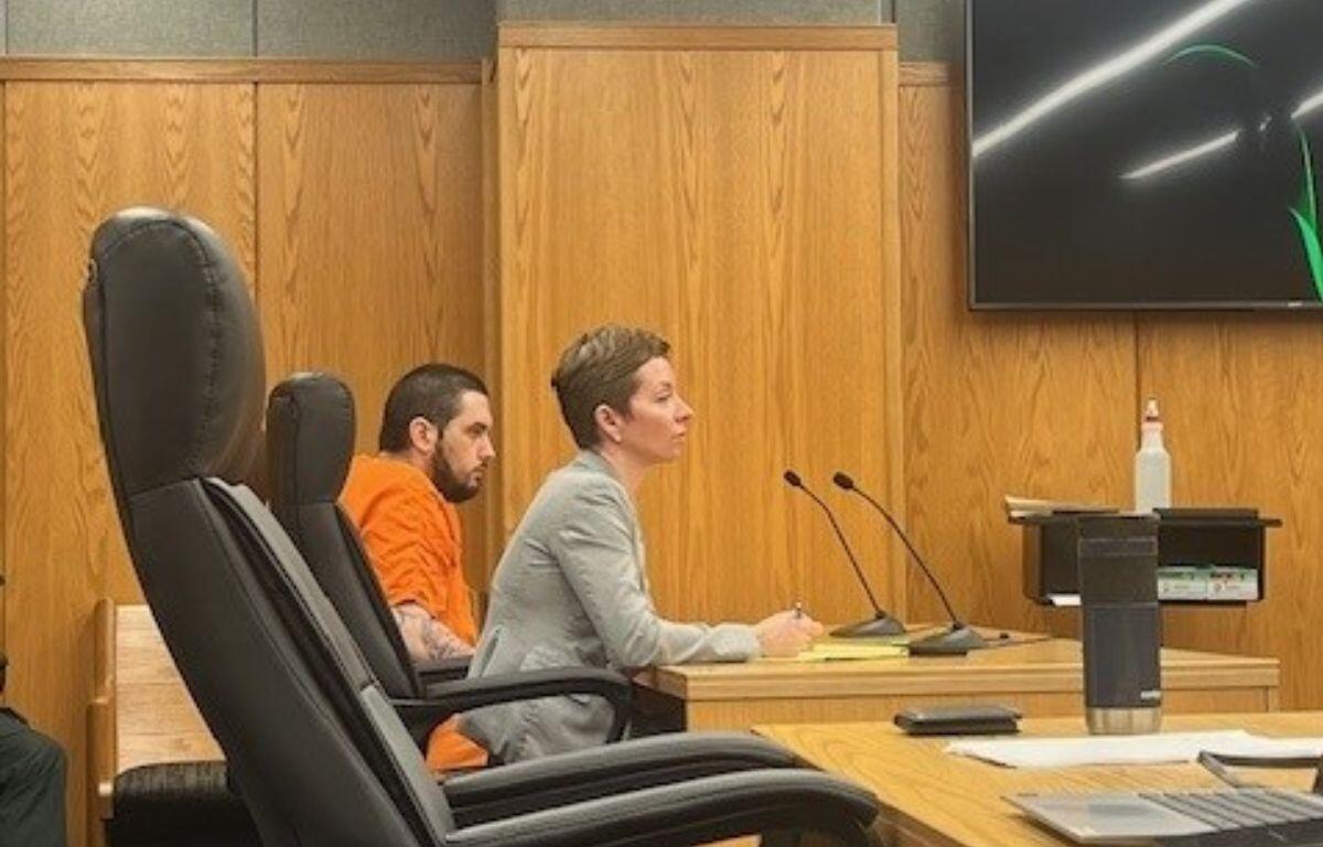 An empty chair in the foreground on the left, a well-dressed woman in the middle of photo and behind her a man in an orange jump suit. They are sitting in front of a long table with microphones in front of them to the right, with wooden wall panels in the background.