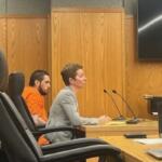 An empty chair in the foreground on the left, a well-dressed woman in the middle of photo and behind her a man in an orange jump suit. They are sitting in front of a long table with microphones in front of them to the right, with wooden wall panels in the background.