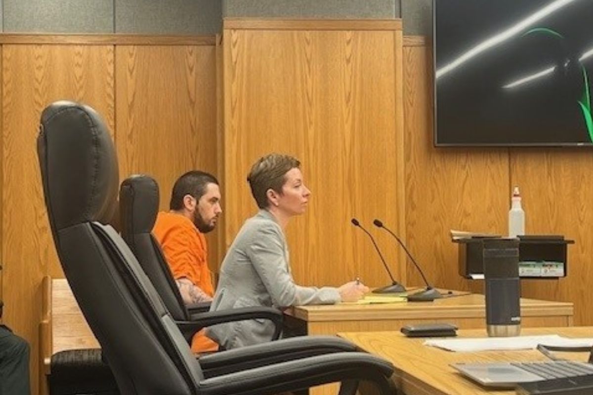 An empty chair in the foreground on the left, a well-dressed woman in the middle of photo and behind her a man in an orange jump suit. They are sitting in front of a long table with microphones in front of them to the right, with wooden wall panels in the background.