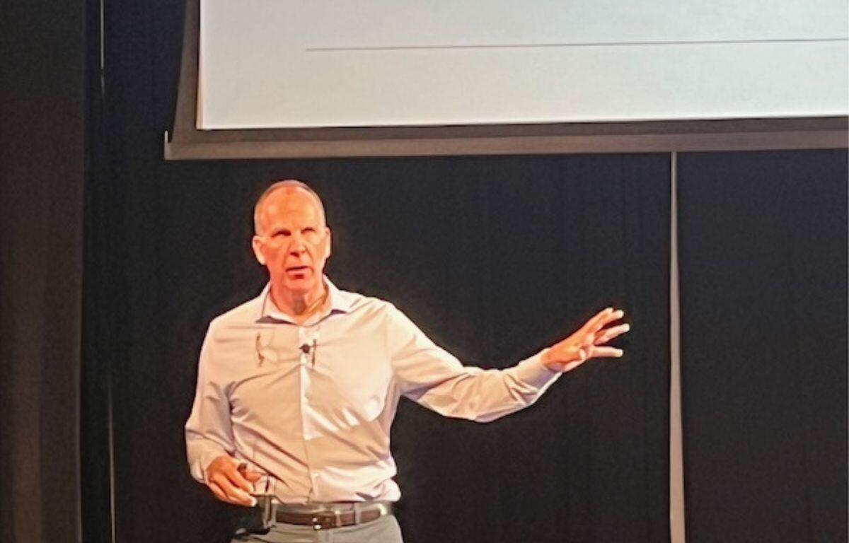 A man standing against a black background, with a monitor overhead, with his left arm extended.