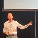 A man standing against a black background, with a monitor overhead, with his left arm extended.