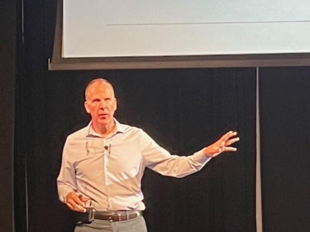 A man standing against a black background, with a monitor overhead, with his left arm extended.