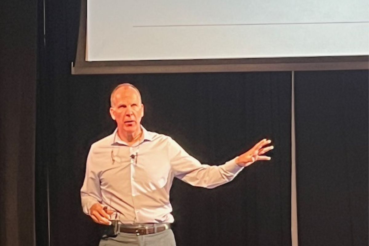 A man standing against a black background, with a monitor overhead, with his left arm extended.
