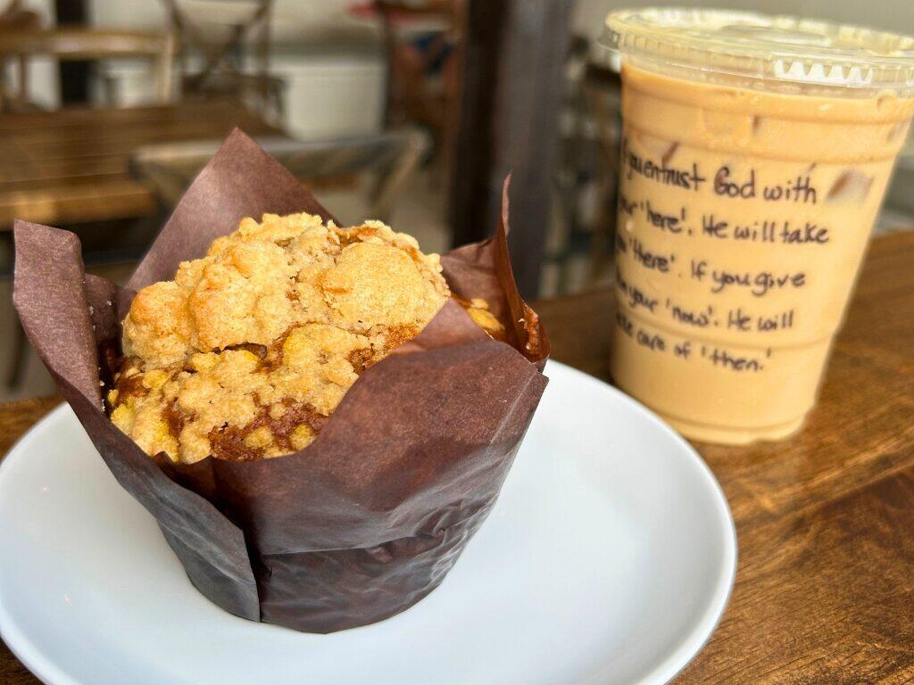 The Gathering Cafe's pumpkin muffin from scratch on a white plate with a pumpkin vanilla latte behind it.