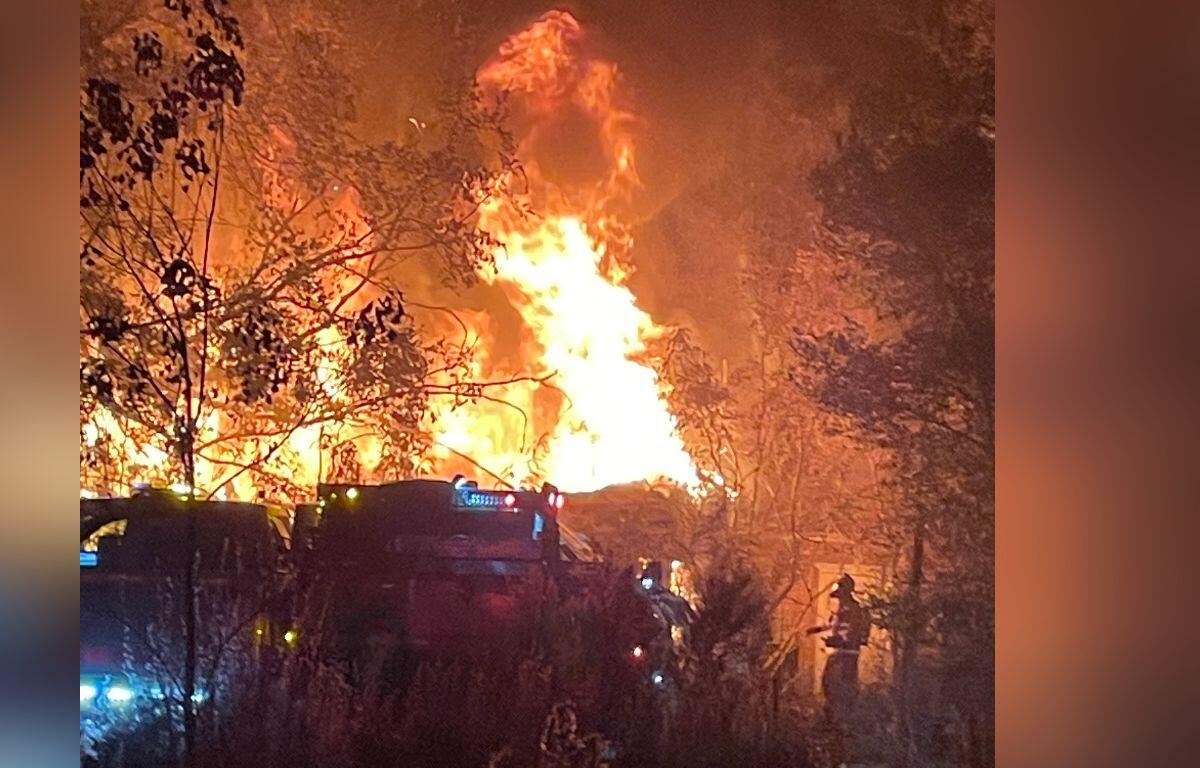 Background shows an illuminated by flames, a tree line to the left, a home fully involved, with a silhouette of fire trucks to the left and fire firefighters to the right.