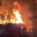 Background shows an illuminated by flames, a tree line to the left, a home fully involved, with a silhouette of fire trucks to the left and fire firefighters to the right.