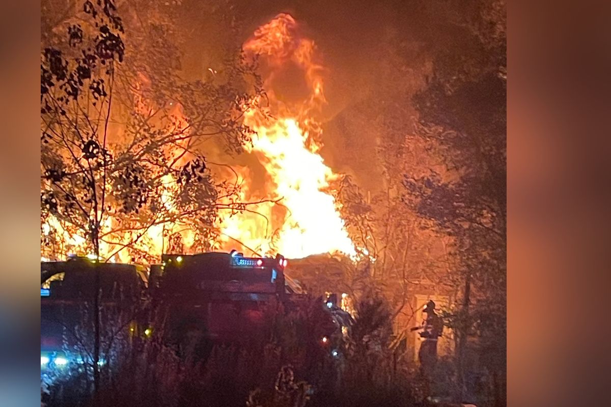 Background shows an illuminated by flames, a tree line to the left, a home fully involved, with a silhouette of fire trucks to the left and fire firefighters to the right.