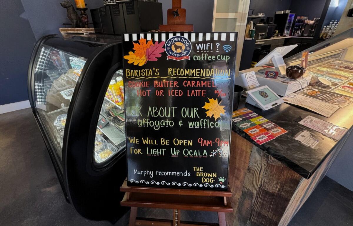 A black easel at Brown Dog Coffee House that has "Barista's Recommendation: Cookie Butter Caramel Hot or Iced Latte 10/10," "Ask about our Affogato & waffleato," "We will be open 9am-9pm for Light up Ocala," and "Murphy recommends The Brown Dog" written on it. Behind the easel there is a pastry case and the register.