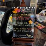 A black easel at Brown Dog Coffee House that has "Barista's Recommendation: Cookie Butter Caramel Hot or Iced Latte 10/10," "Ask about our Affogato & waffleato," "We will be open 9am-9pm for Light up Ocala," and "Murphy recommends The Brown Dog" written on it. Behind the easel there is a pastry case and the register.