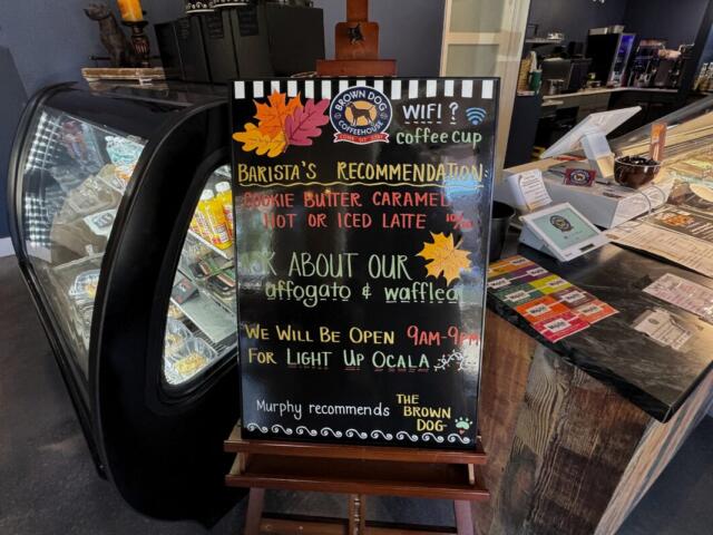 A black easel at Brown Dog Coffee House that has "Barista's Recommendation: Cookie Butter Caramel Hot or Iced Latte 10/10," "Ask about our Affogato & waffleato," "We will be open 9am-9pm for Light up Ocala," and "Murphy recommends The Brown Dog" written on it. Behind the easel there is a pastry case and the register.
