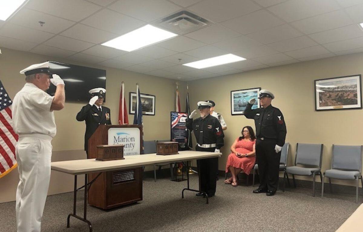 On the extreme left the U.S. flag, a man dressed in uniform saluting, in background a monitor hanging on the wall, a man in uniform behind a podium, that's behind a table with two wooden urns, two flags in front of a framed picture, a placard with writing in front of two flags, two other men in uniform saluting, another man in uniform against the wall, two more framed pictures on the right, a woman in chair, and empty chairs to her right.