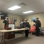 On the extreme left the U.S. flag, a man dressed in uniform saluting, in background a monitor hanging on the wall, a man in uniform behind a podium, that's behind a table with two wooden urns, two flags in front of a framed picture, a placard with writing in front of two flags, two other men in uniform saluting, another man in uniform against the wall, two more framed pictures on the right, a woman in chair, and empty chairs to her right.