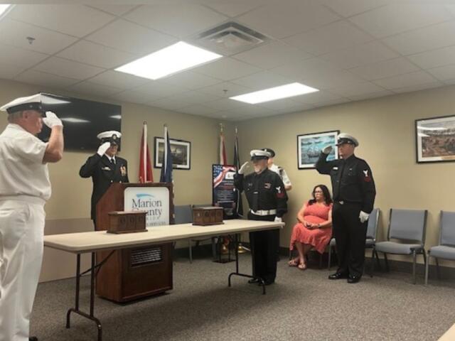On the extreme left the U.S. flag, a man dressed in uniform saluting, in background a monitor hanging on the wall, a man in uniform behind a podium, that's behind a table with two wooden urns, two flags in front of a framed picture, a placard with writing in front of two flags, two other men in uniform saluting, another man in uniform against the wall, two more framed pictures on the right, a woman in chair, and empty chairs to her right.