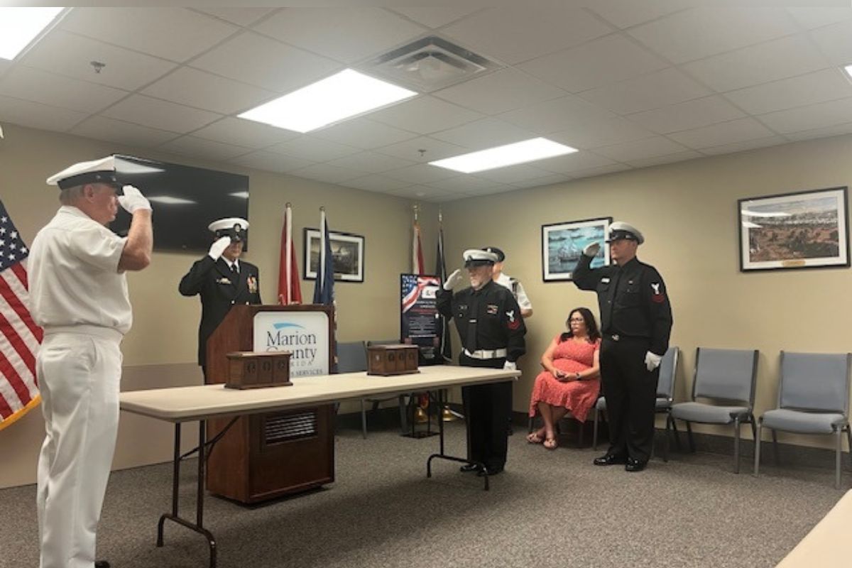 On the extreme left the U.S. flag, a man dressed in uniform saluting, in background a monitor hanging on the wall, a man in uniform behind a podium, that's behind a table with two wooden urns, two flags in front of a framed picture, a placard with writing in front of two flags, two other men in uniform saluting, another man in uniform against the wall, two more framed pictures on the right, a woman in chair, and empty chairs to her right.