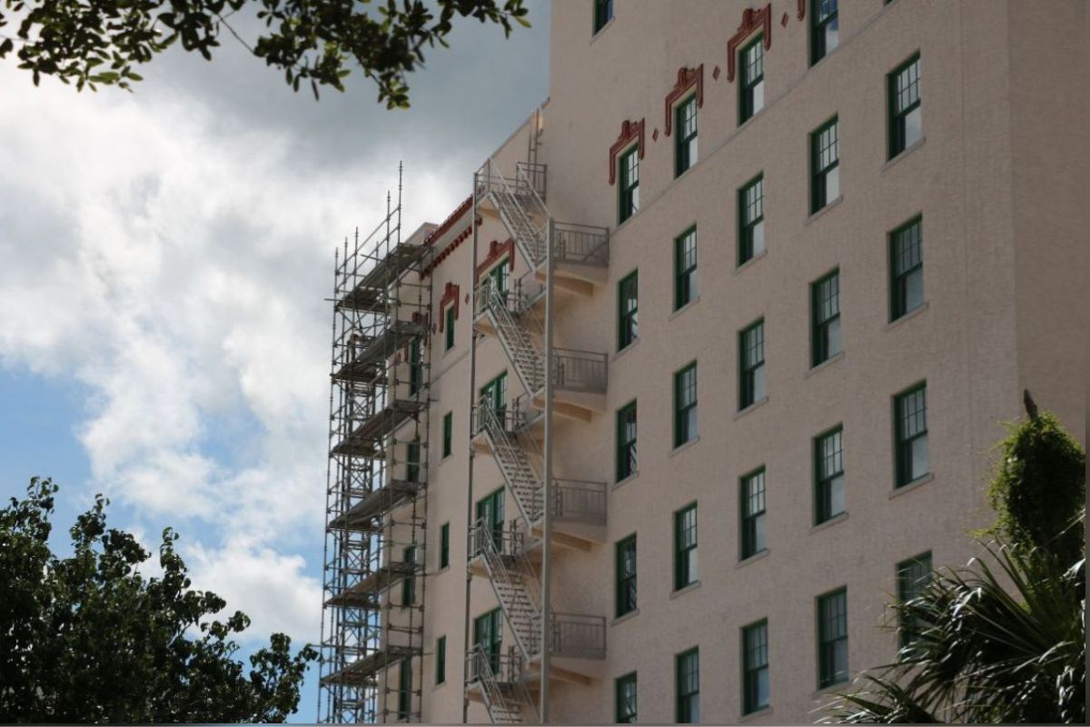 Blue skies and white clouds in the background, a building with scaffolding to the extreme left, and to the right of that a fire escape, and in the foreground, shade trees on the left and an ornamental palm on the right.