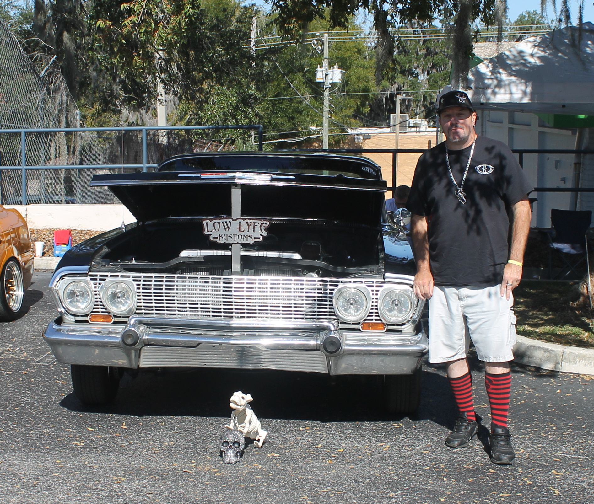A skull and a skeleton in front of a vintage classic car, with a man in a baseball hat, t-shirt, shorts and red socks to the right of the car, with trees and poles in the background and pavement in the foreground. 