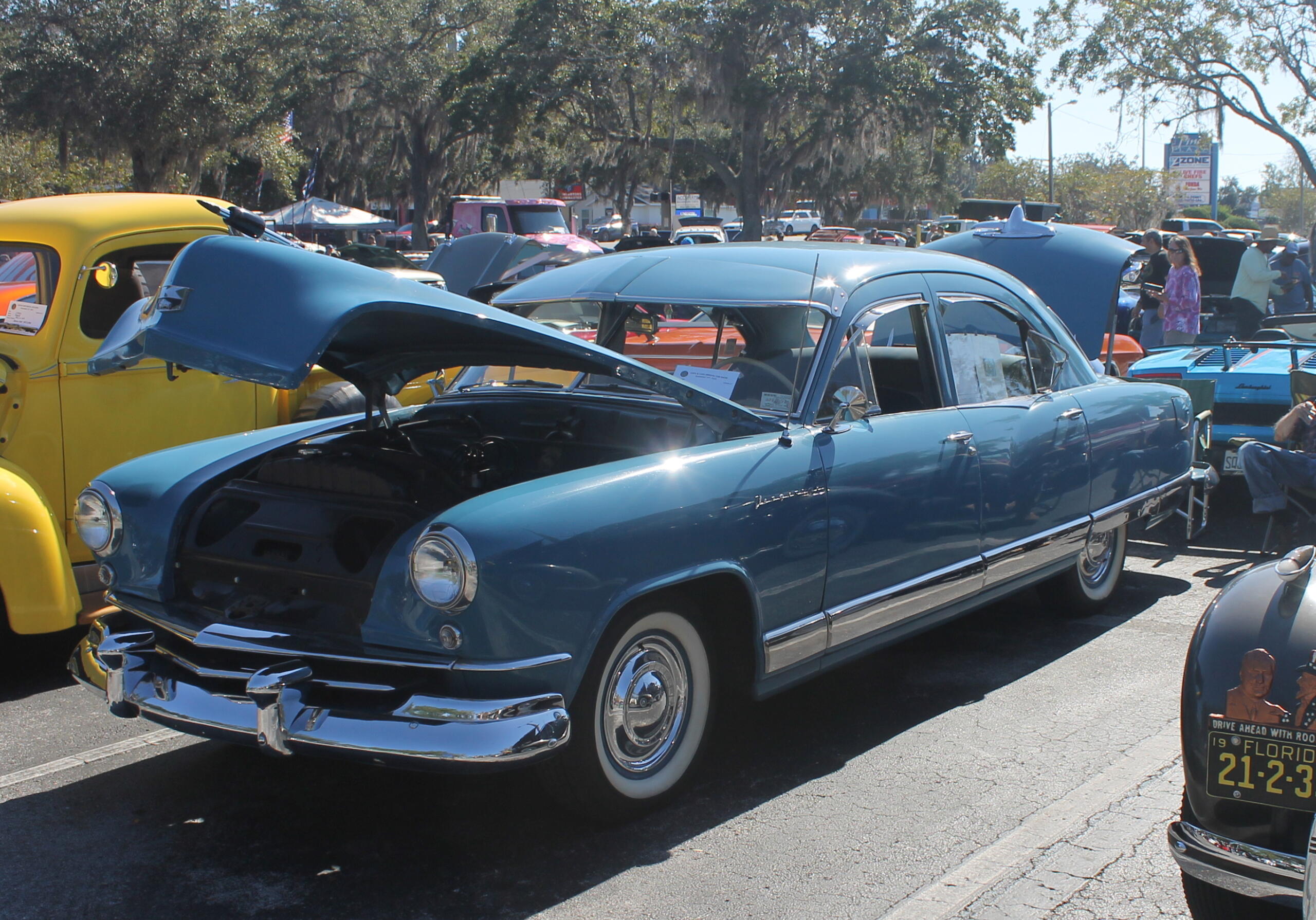 A blue vehicle with its hood open, with a vehicle to the left and pavement to the right, with trees in the background.