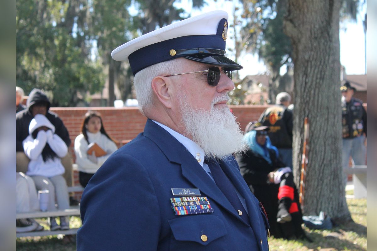 A man dressed in a military uniform, with people in the background standing in front of a brick wall and trees.