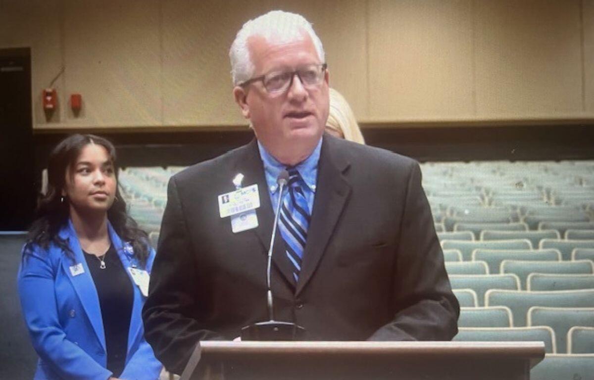 A woman in a blue blazer standing in the background to the left, a fire alarm on the far beige wall with brown trim on the back. A sea of empty gray suits, and a woman with blonde hair standing behind the speaker, wearing a black blazer who is in front of a podium with a microphone.