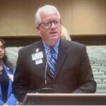 A woman in a blue blazer standing in the background to the left, a fire alarm on the far beige wall with brown trim on the back. A sea of empty gray suits, and a woman with blonde hair standing behind the speaker, wearing a black blazer who is in front of a podium with a microphone.