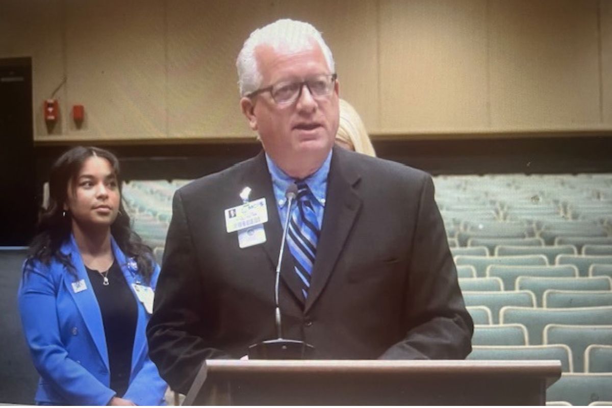 A woman in a blue blazer standing in the background to the left, a fire alarm on the far beige wall with brown trim on the back. A sea of empty gray suits, and a woman with blonde hair standing behind the speaker, wearing a black blazer who is in front of a podium with a microphone.