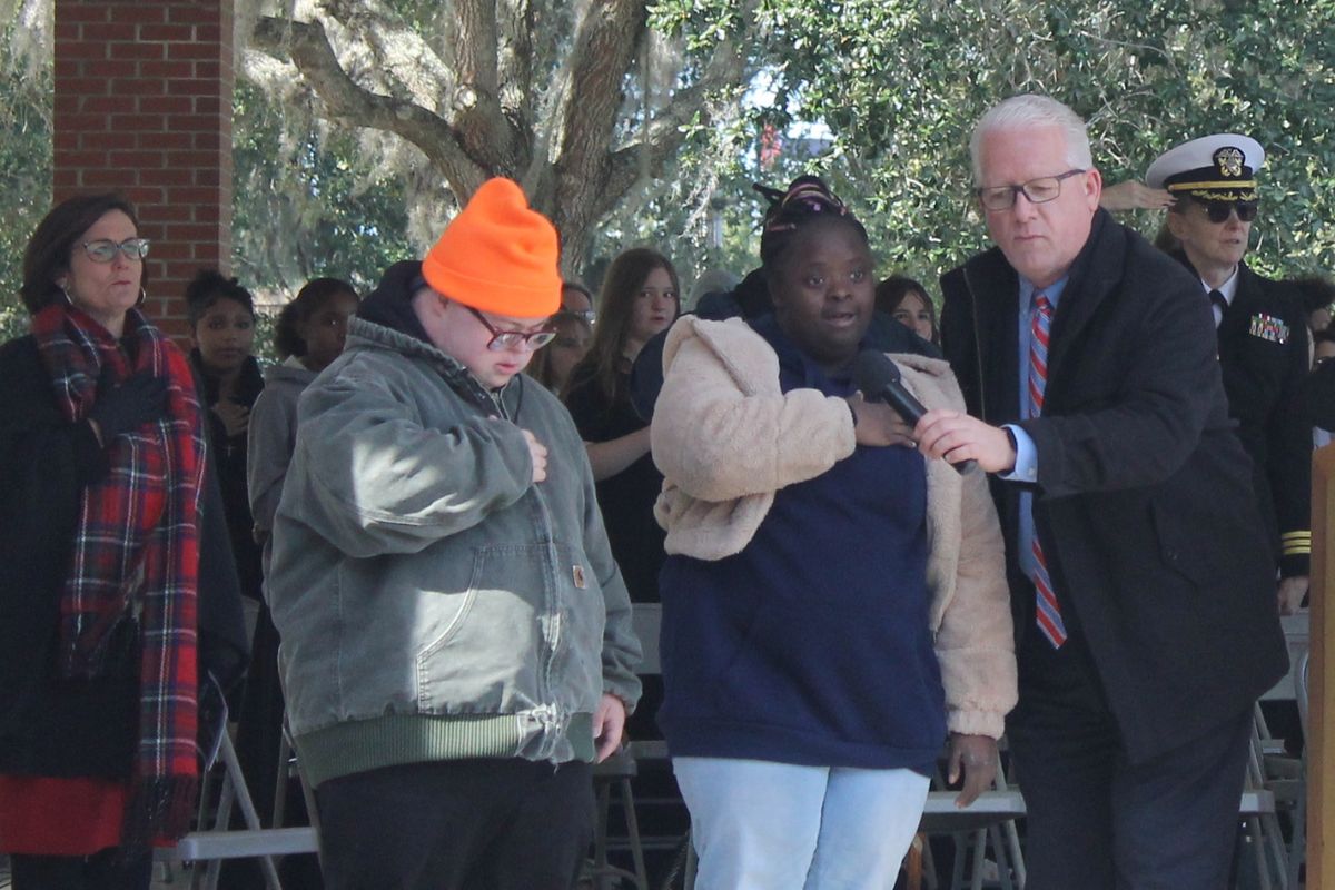 A woman wearing glasses, and overcoat and scarf, with her hand over her chest, a man wearing a hat and overcoat with his hand over his chest, a woman wearing an overcoat with a hand over her chest and a man in a suit holding a microphone.