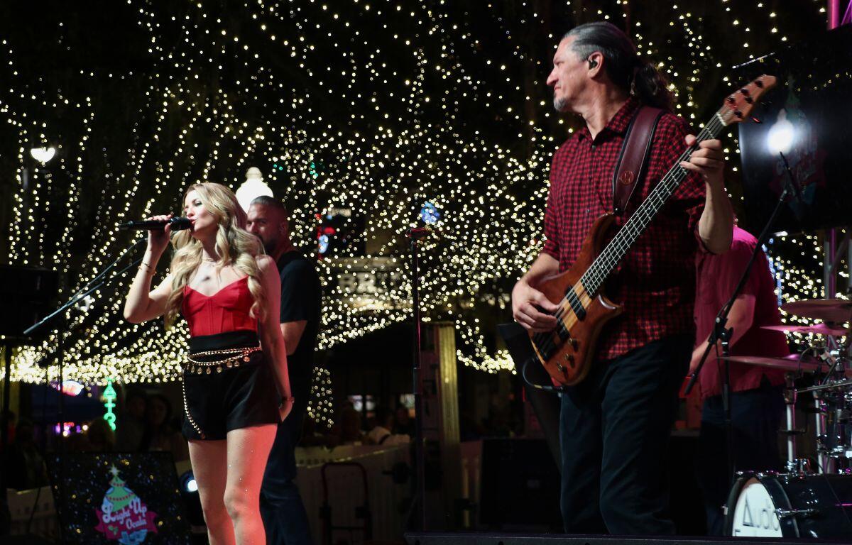Two musicians, a man in a red and black flannel shirt and jeans holding a guitar and a woman wearing a red tank top and black shorts with a gold belt, perform under a display of Christmas lights.