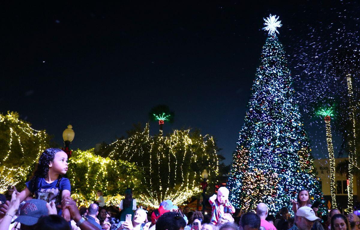 A crowd of people gather around a brightly lit Christmas tree and light display in a town square.
