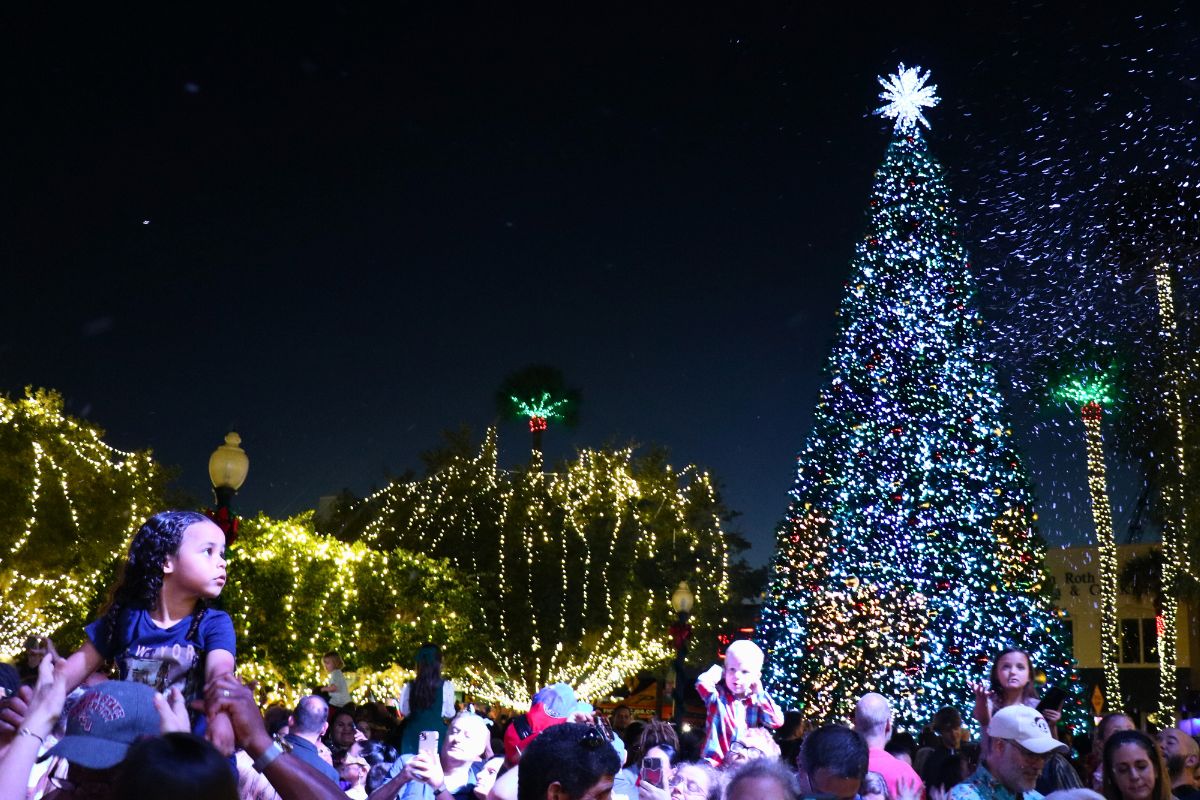 A crowd of people gather around a brightly lit Christmas tree and light display in a town square.