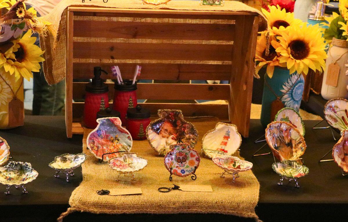 A vendor booth displays hand-painted seashells on a burlap table runner with sunflowers in decorative vases.