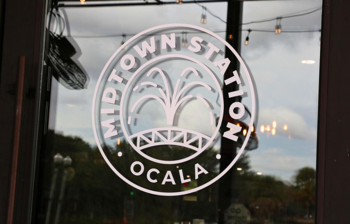 A white logo on a glass door has a fountain in the center with words in a circle around it reading, "Midtown Station Ocala."